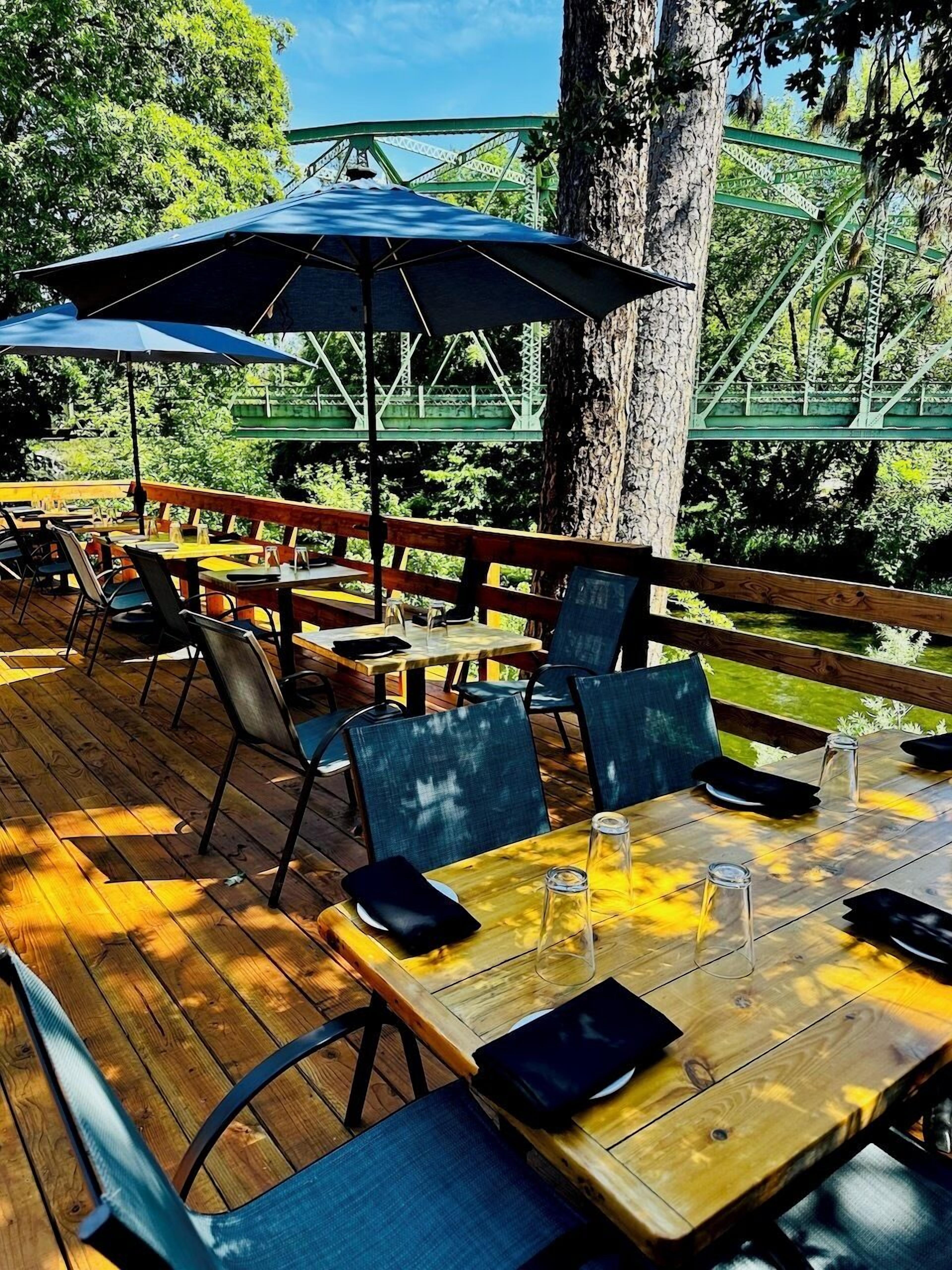 Un espacio de comedor al aire libre con mesas y sillas de madera, un paraguas grande y vista a un puente verde.