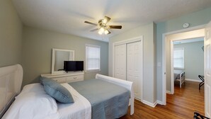 Un dormitorio con cama, cómoda, televisor, ventilador de techo y puerta que da a otra habitación.