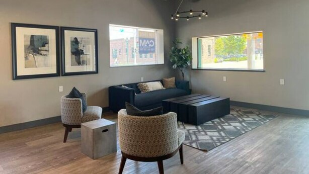 A modern living room with a dark blue sofa, two patterned armchairs, a coffee table, and a large window with a view of a building outside. - Studio63 on Phillips (Sioux Falls)