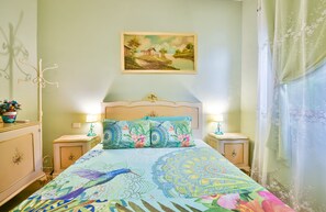 Une chambre à coucher avec un lit, deux tables de chevet, une commode, une lampe, un tableau au mur et une fenêtre avec des rideaux.
