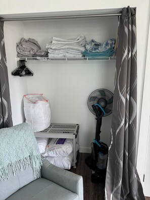 A compact living space with a grey sofa, a small table with a white bag, a standing fan, and a ceiling fan.