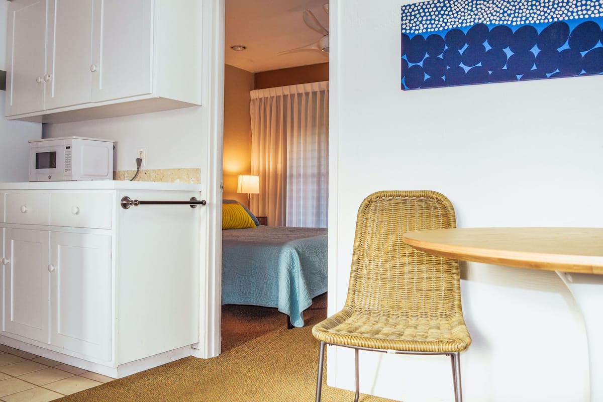 Een compacte hotelkamer met kitchenette, een bed met blauw beddegoed, een rieten stoel en een klein houten tafeltje.