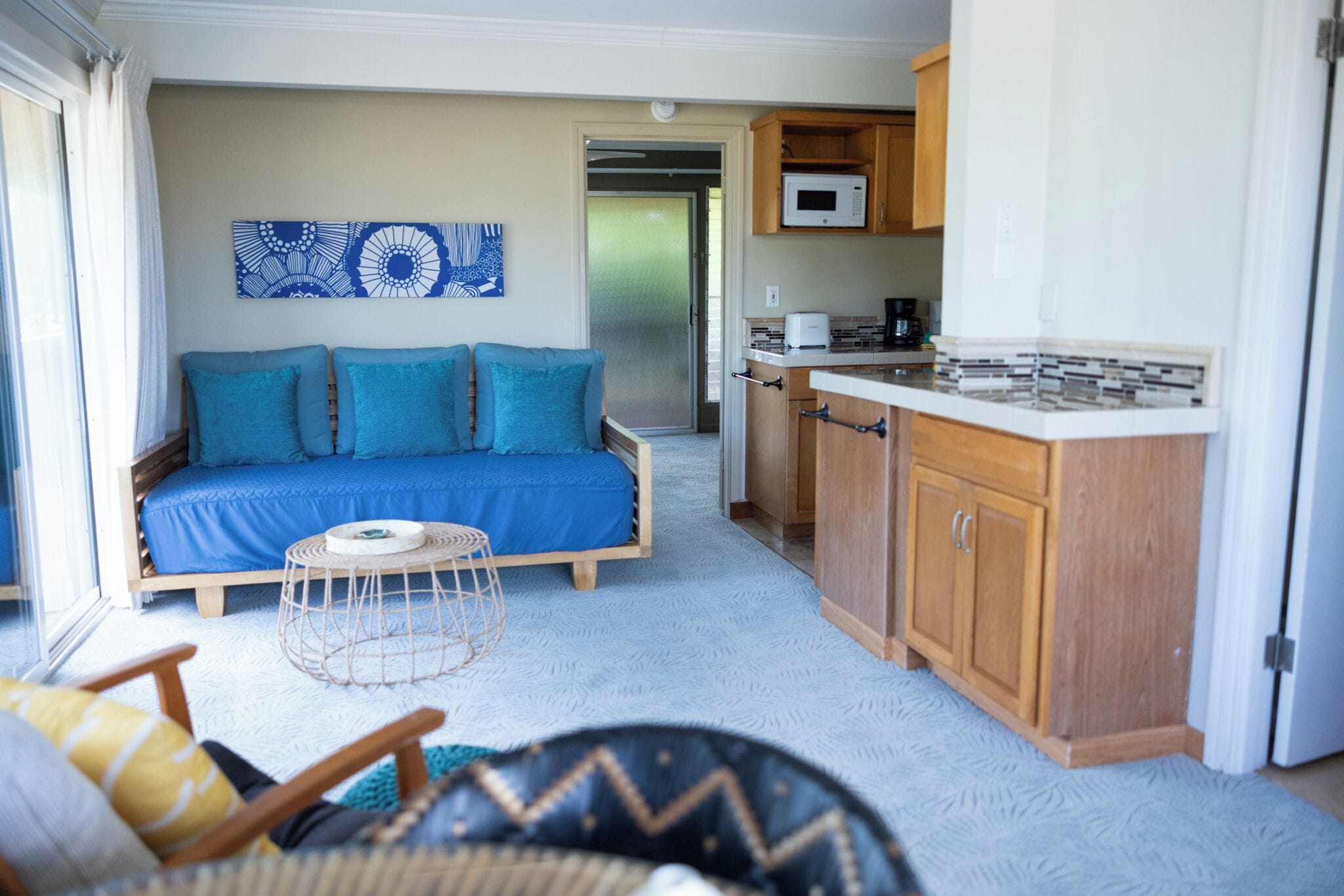 A living room with a blue sofa, a small round table, and a kitchen area with wooden cabinets and a microwave.