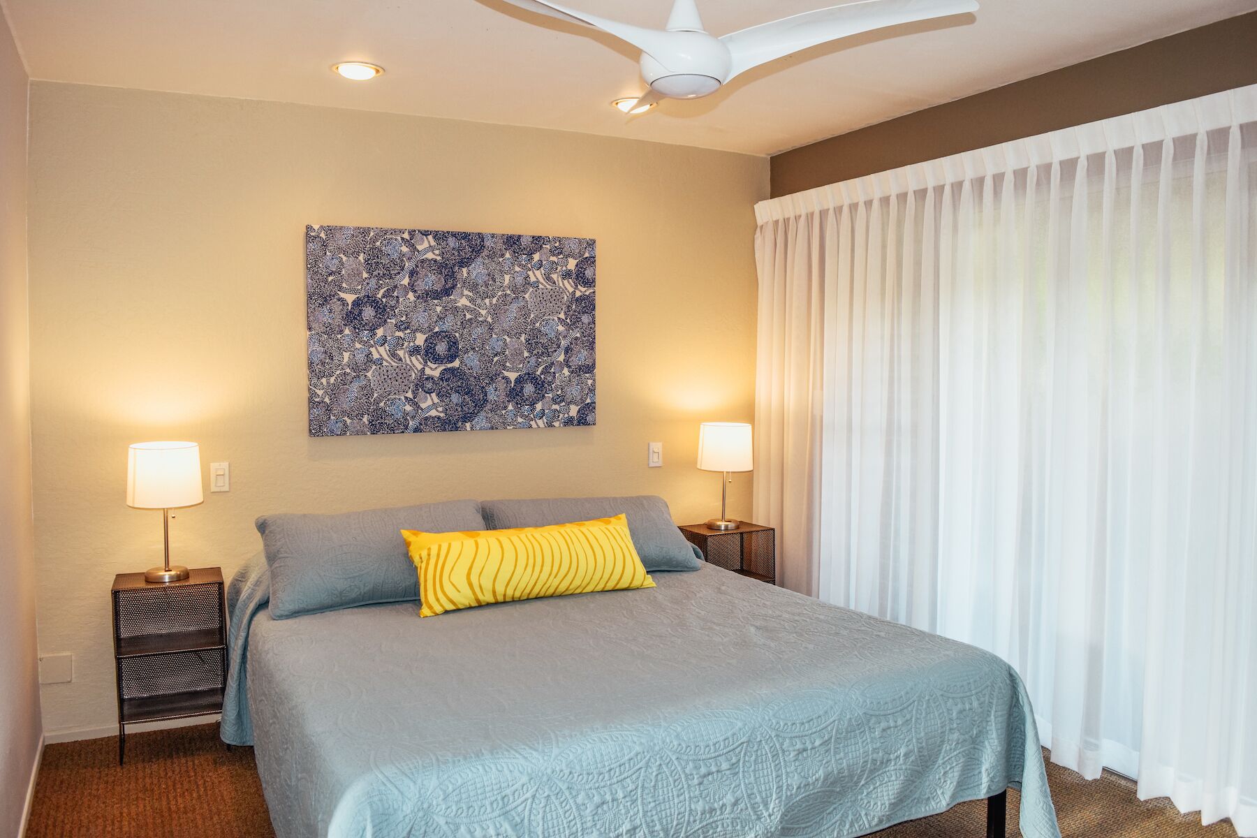 A hotel room with a bed, two bedside lamps, a ceiling fan, a large abstract painting, and sheer curtains.