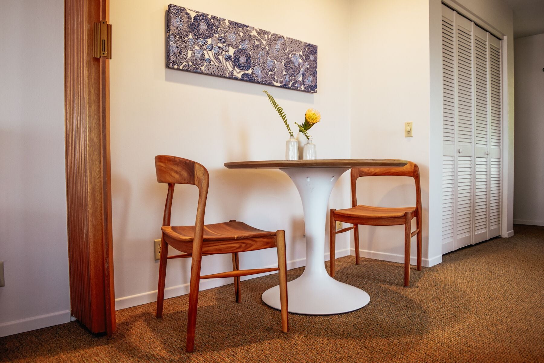 Une pièce avec une table ronde blanche, deux chaises en bois, une œuvre d’art abstraite accrochée au mur et une porte blanche dotée de volets à persiennes.