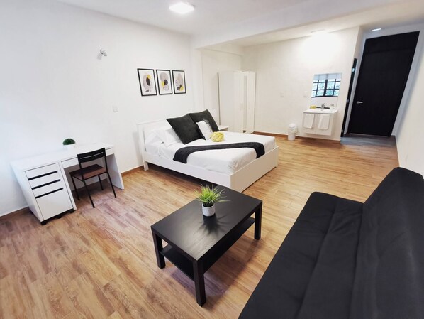 A modern, well-lit room with a bed, a black sofa, a small desk, and a potted plant.