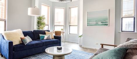 A living room with a blue sofa, a round coffee table, and a view of the city through large windows.