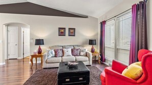 Un salon avec un canapé, un fauteuil rouge, une table basse et une grande fenêtre avec des rideaux.