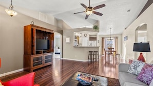 Un salon spacieux avec un meuble de rangement en bois pour les appareils multimédias, un ventilateur de plafond et un coin repas à l’arrière-plan.