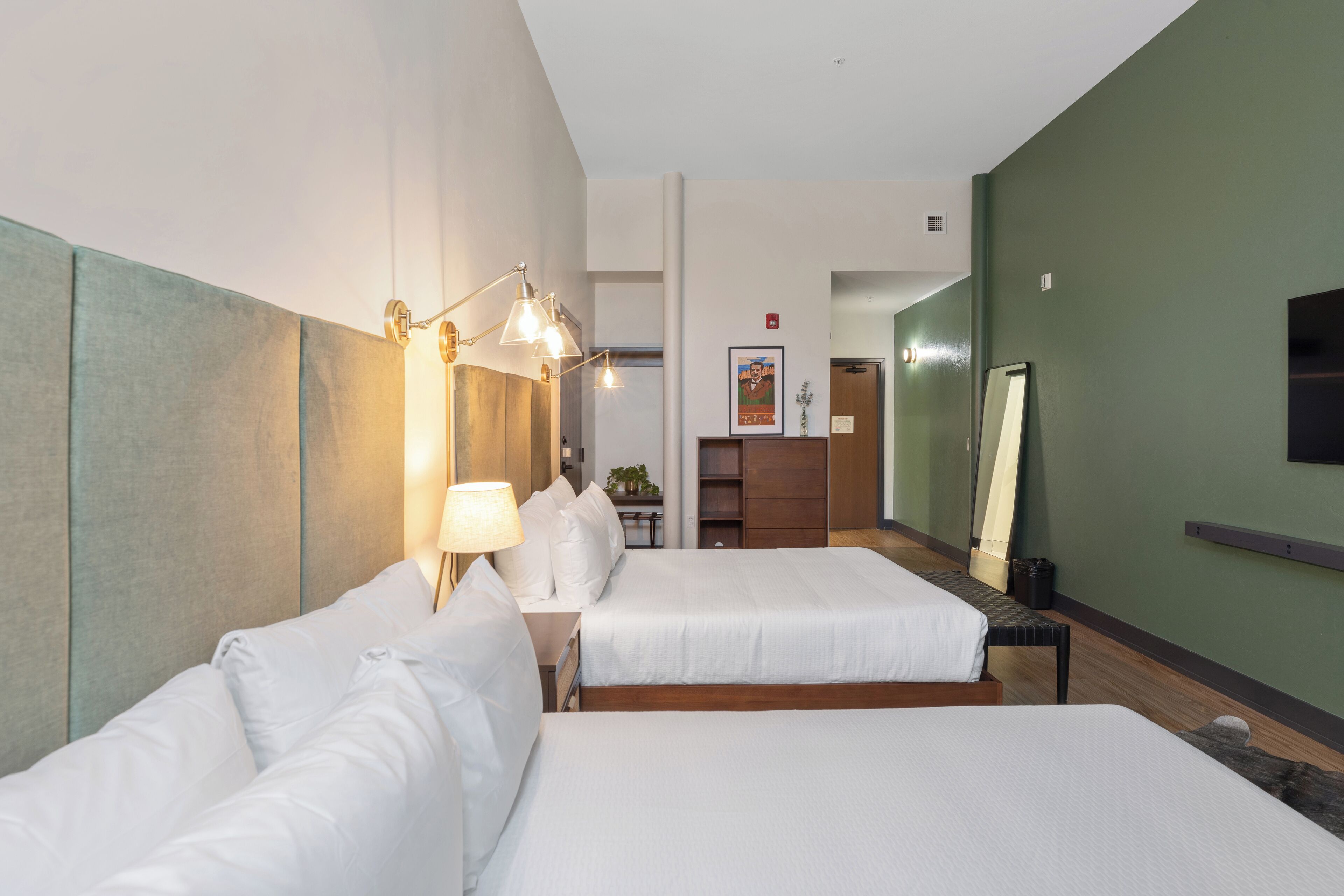 A hotel room with two beds, a desk, a chair, a lamp, a mirror, and a small plant.