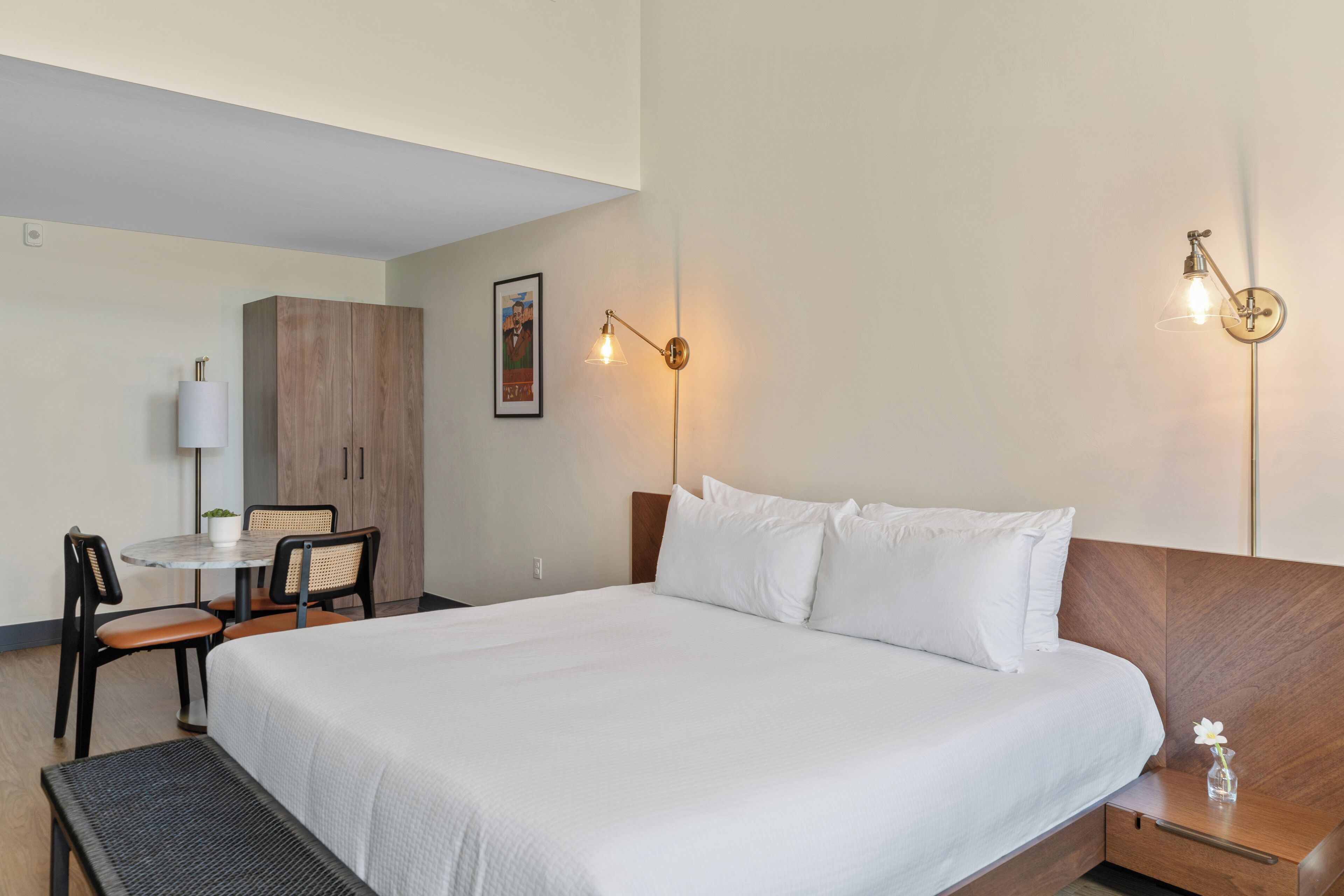 A hotel room with a large bed, a desk with chairs, a lamp, and a wall-mounted picture.