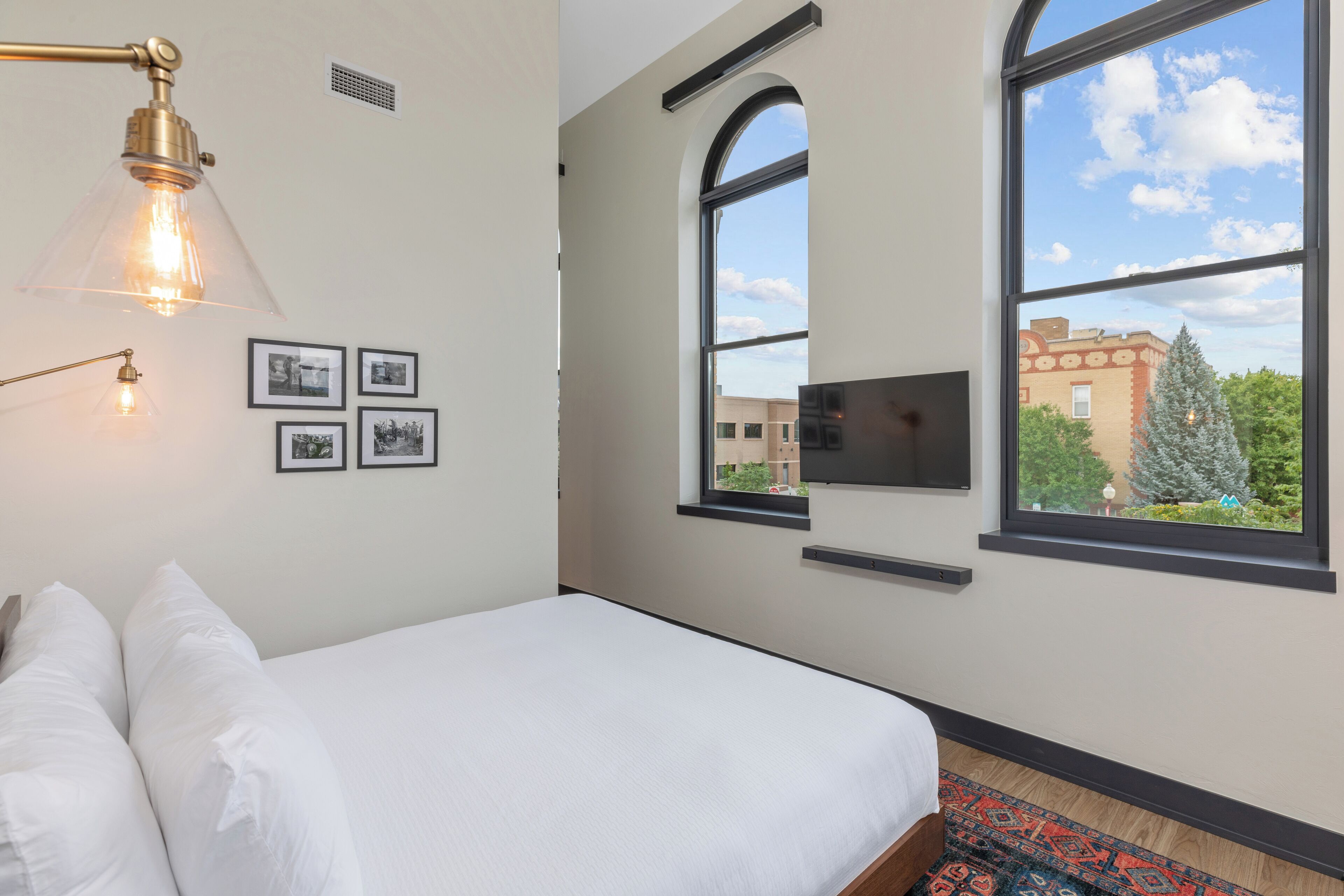A bedroom with a bed, a wall-mounted TV, and a window with a view of buildings and trees.