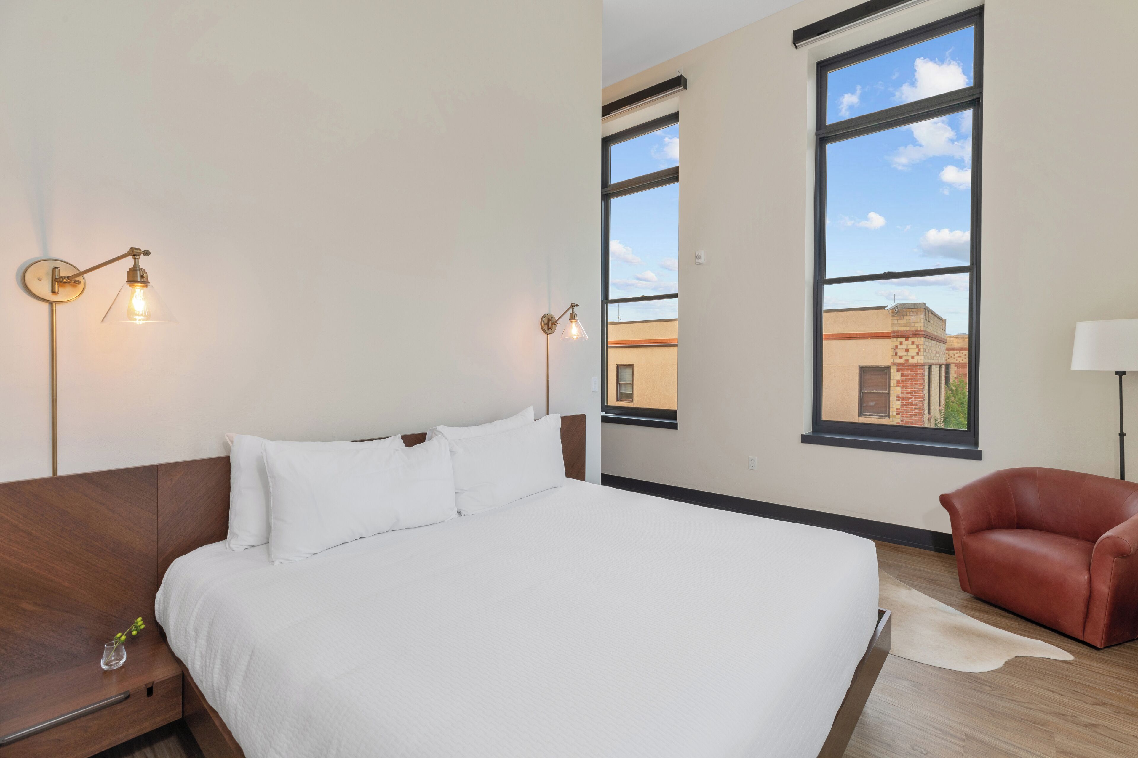 A modern hotel room with a large bed, two wall-mounted lights, a leather chair, and a view of the city through large windows.