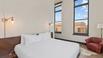 A modern hotel room with a large bed, two wall-mounted lights, a leather chair, and a view of the city through large windows.