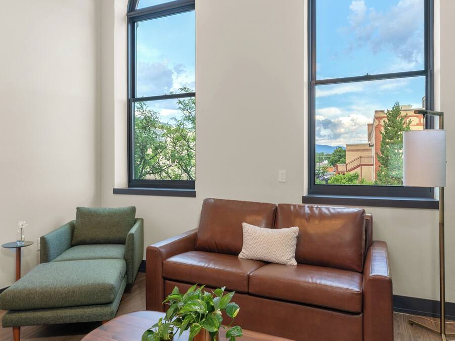 A modern living room with a brown leather sofa, a green armchair, and a wooden coffee table with a plant. There are two large arched windows providing views of buildings and trees.