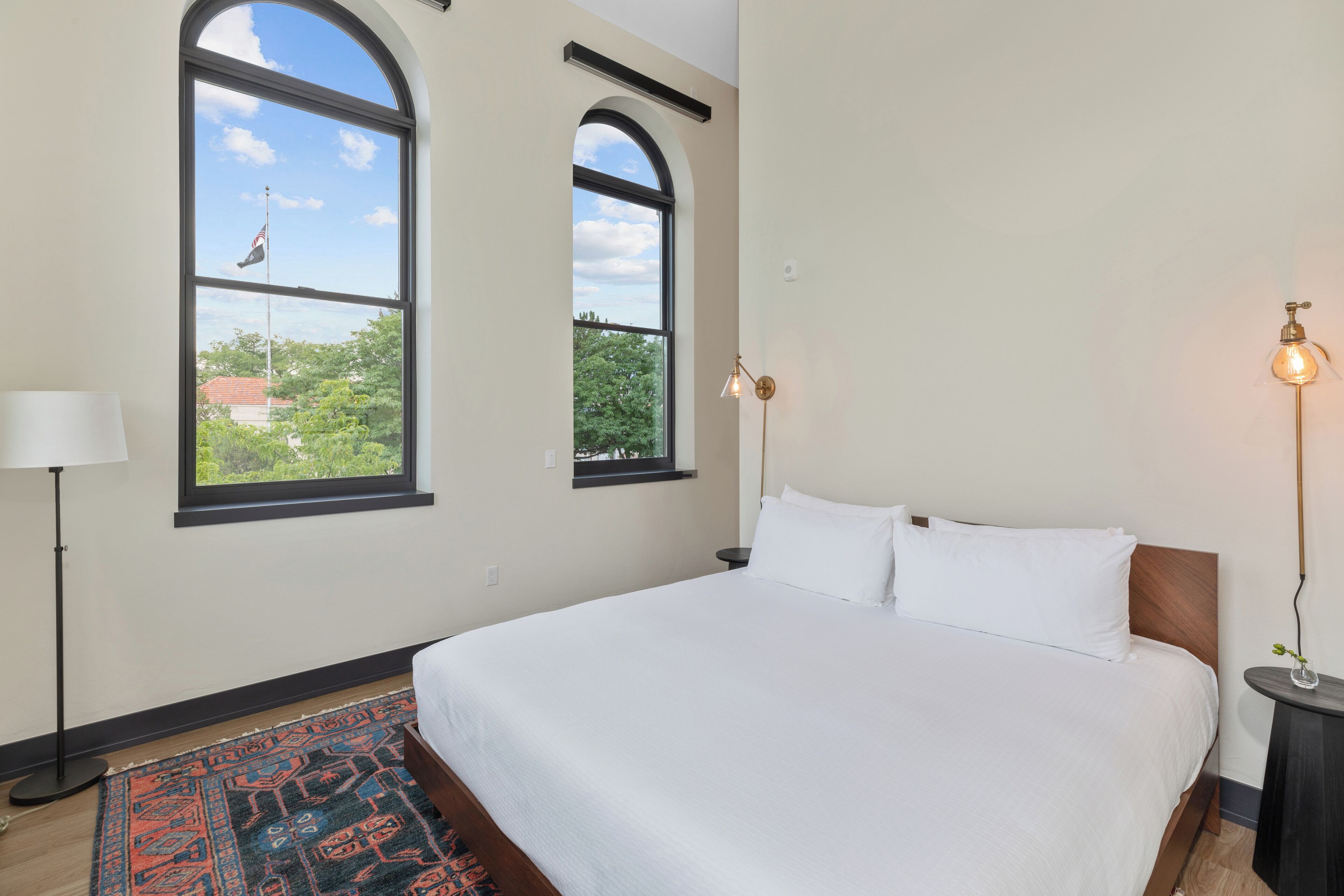 A bedroom with a large bed, two windows, a nightstand, a lamp, and a rug.
