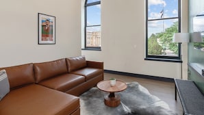 A modern living room with a brown leather sofa, a round wooden coffee table with a plant, and large windows offering a view of buildings and trees. - The Rathbone Hotel (Montrose)