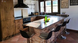 A modern kitchen with wooden cabinets, a large island with bar stools, a built-in oven, and a window with a view of greenery.