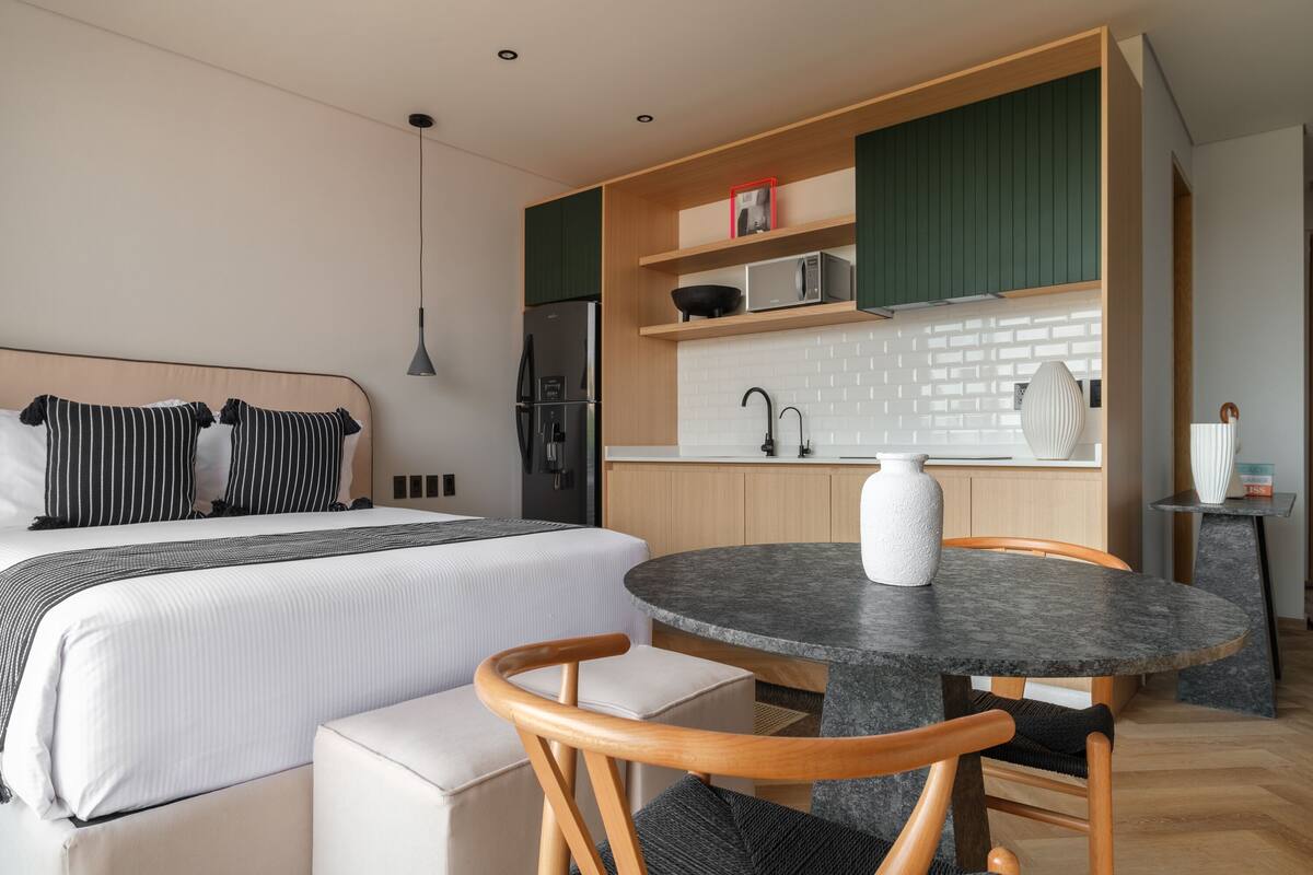 A modern hotel room with a bed, a round dining table, wooden chairs, a kitchen area with a microwave, and a small cabinet.