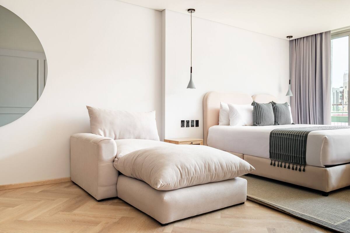 A modern hotel room with a sofa, a bed with a striped blanket, and a view of the city.