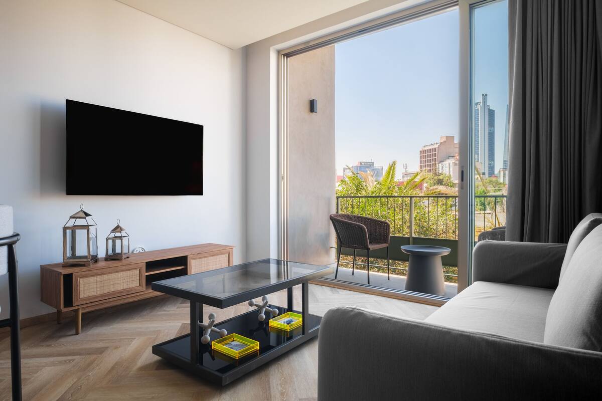 A modern living room with a flat-screen TV, a glass coffee table with decorative items, a sofa, and a view of the city through large windows.