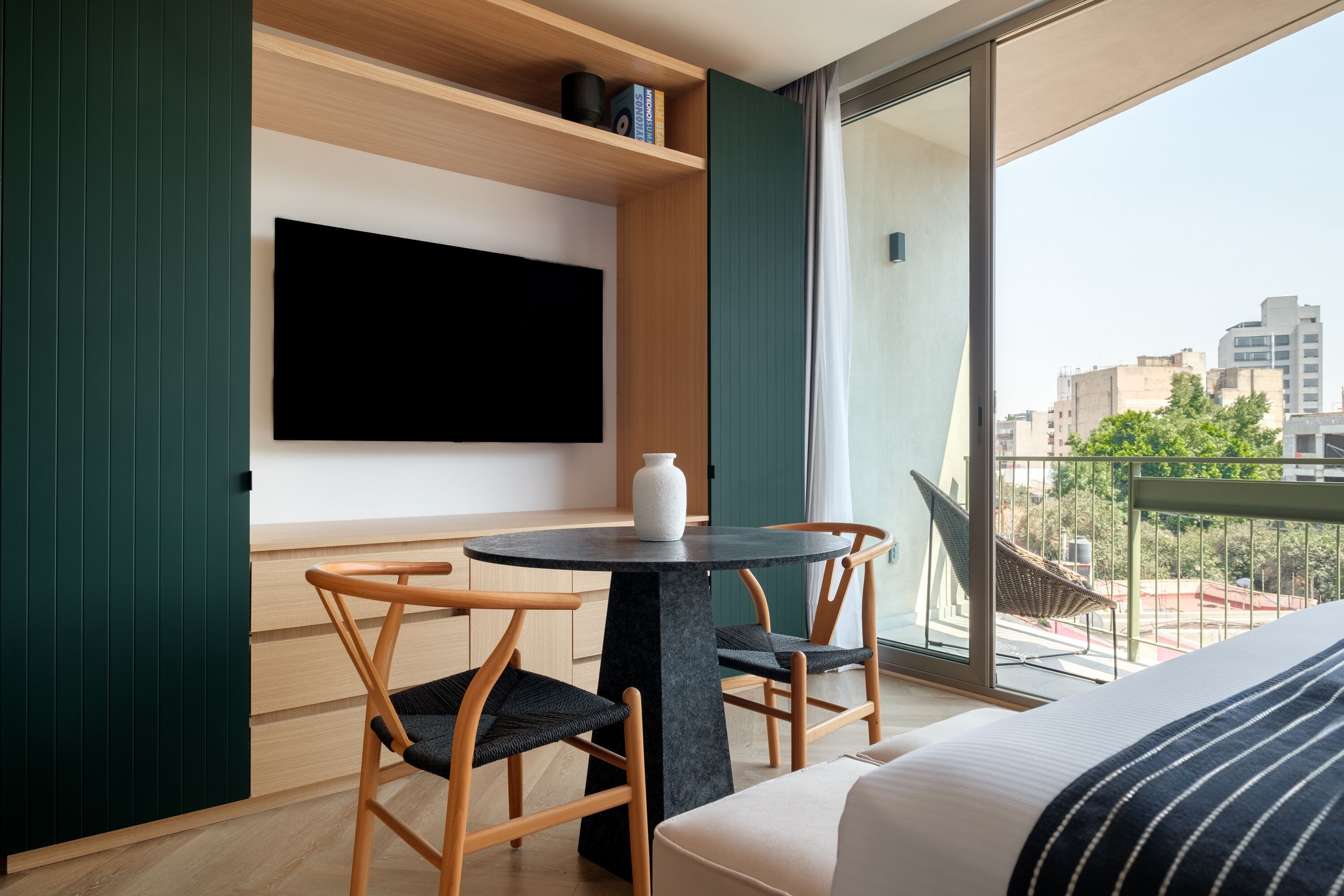 A modern hotel room with a large bed, a round table with two chairs, a flat-screen TV mounted on the wall, and a balcony with a view of the city.
