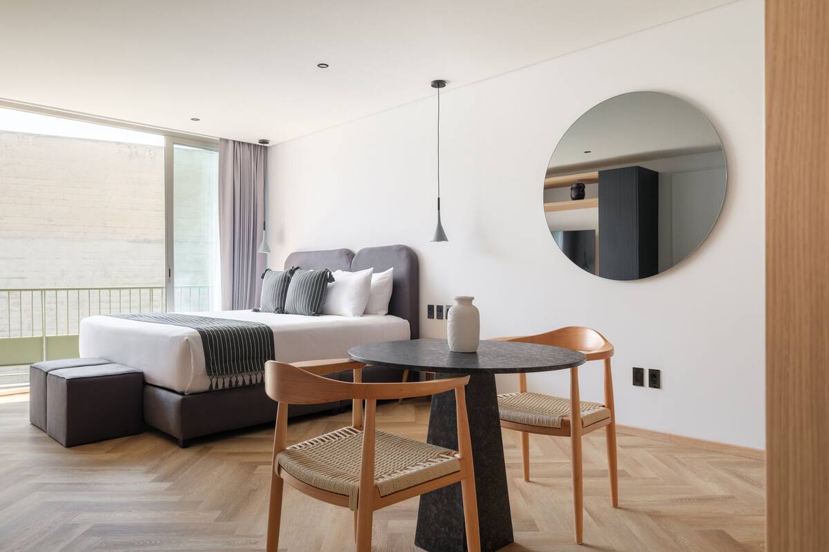 A modern hotel room with a bed, a round table, two chairs, a large mirror, and a view of the outside.