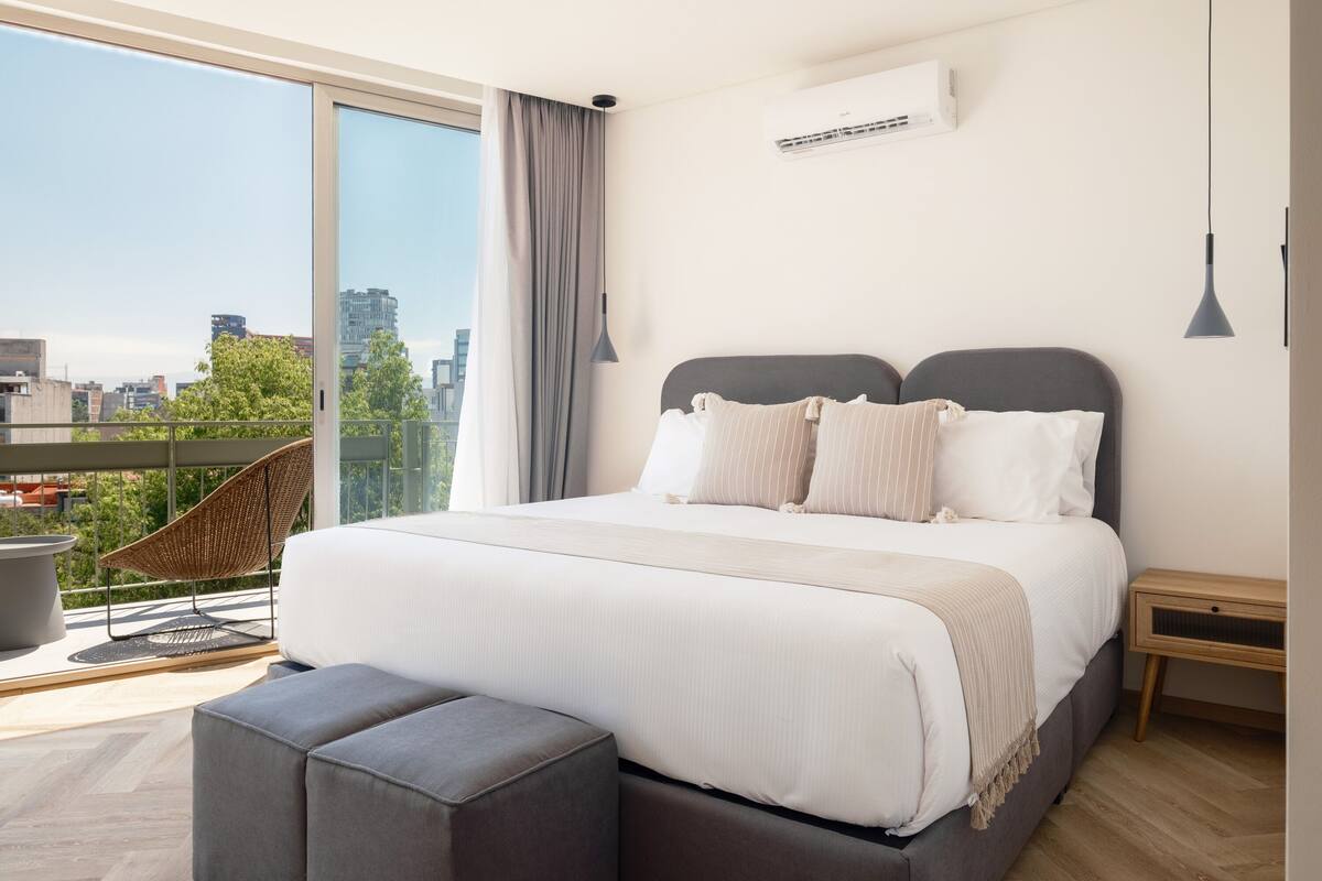 A modern bedroom with a large bed, a balcony with a wicker chair, and a city view.