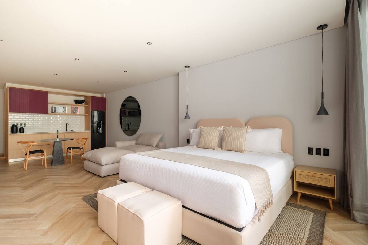 A modern bedroom with a large bed, a small desk, and a kitchenette in the background.