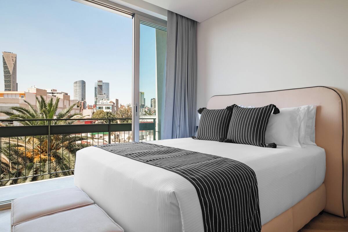 A modern hotel room with a large bed, a view of the cityscape, and a balcony with palm trees.