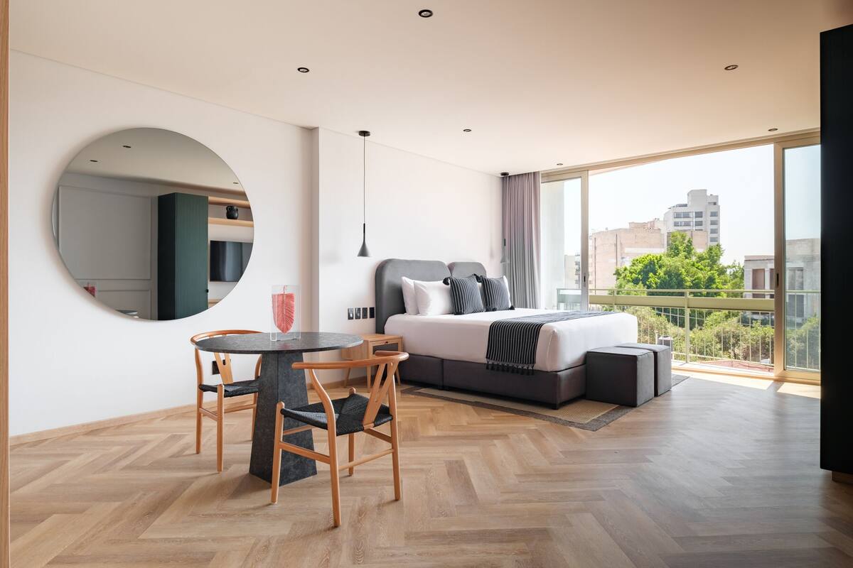 A modern hotel room with a large bed, a round dining table, and a view of the city through large windows.