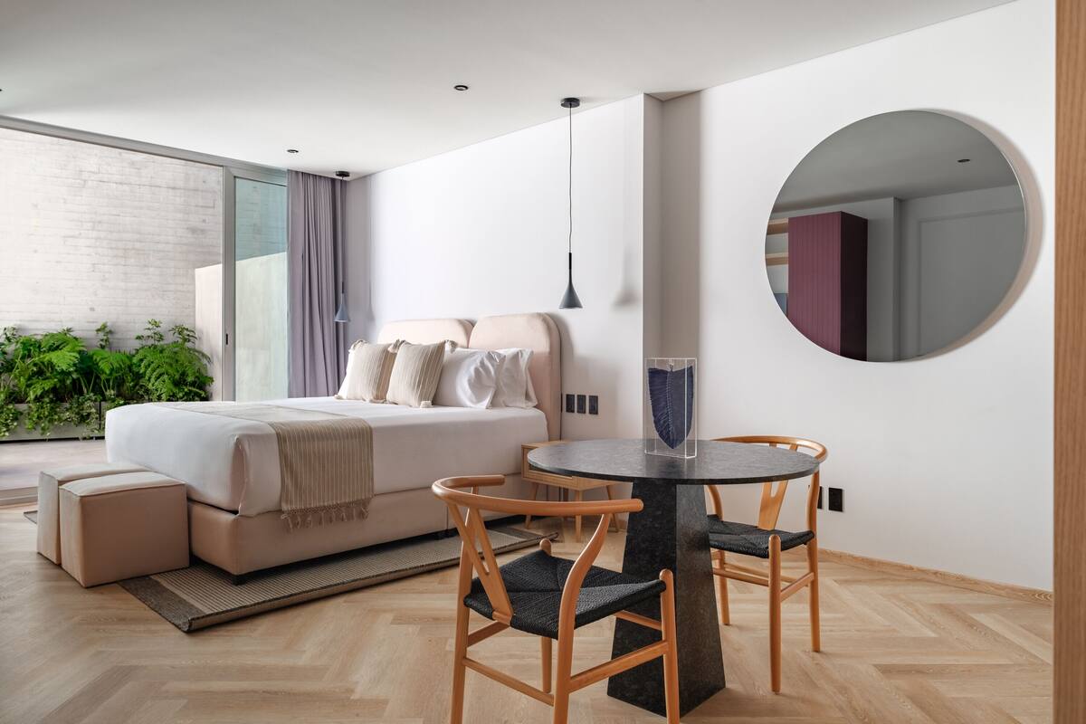 A modern bedroom with a bed, bedside tables, a round dining table with chairs, a large mirror, and a view of the outdoors.