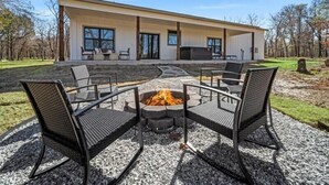 Un patio moderno al aire libre con un brasero, rodeado de sillas de mimbre negro, y un área de estar cubierta con vista a una casa grande al fondo.
