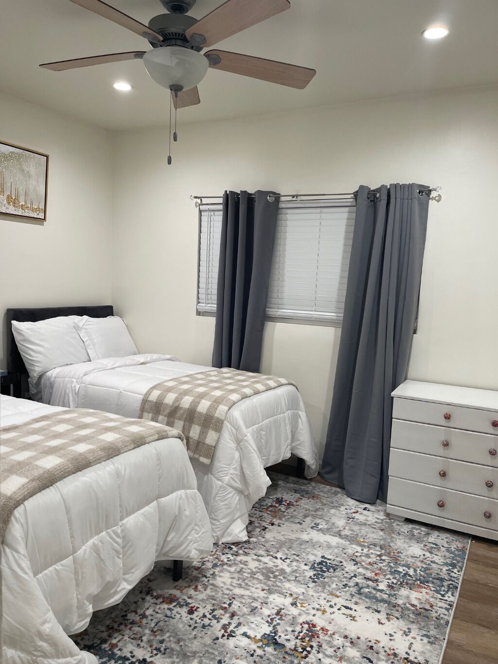 A bedroom with two beds, a ceiling fan, a window with curtains, a dresser, and a patterned rug.