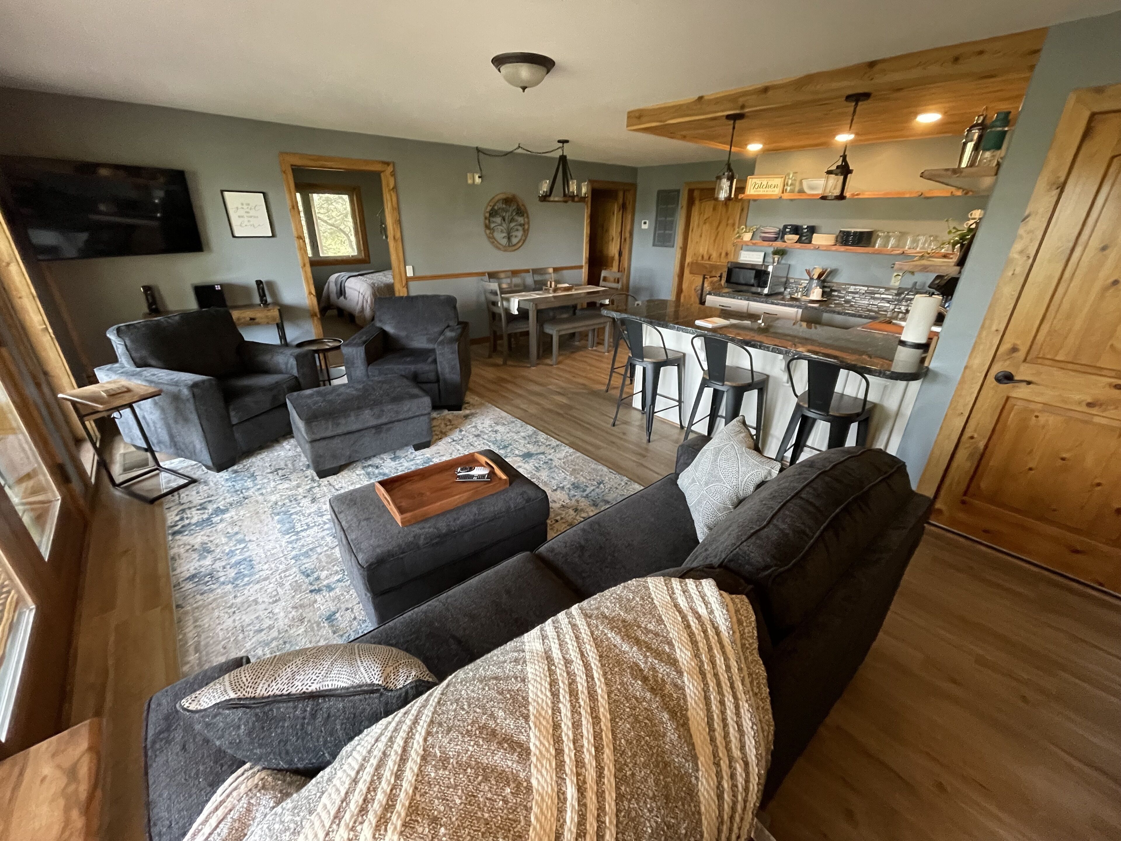 Un salon moderne avec un canapé gris, un pouf et une table basse. On y trouve également un coin cuisine avec des meubles en bois et une table à manger avec des chaises.