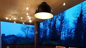 An outdoor wooden deck with a table and chairs, a hanging pendant light, and a view of trees and a house in the background.