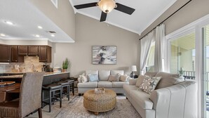 A spacious living room with a large sofa, a coffee table, and a dining area with a kitchen in the background. - Sandy's Beach Retreat (Miramar Beach)