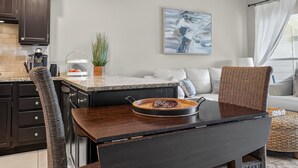 A modern kitchen with dark cabinetry, a granite countertop, and a dining area with a dark table and wicker chairs. - Sandy's Beach Retreat (Miramar Beach)