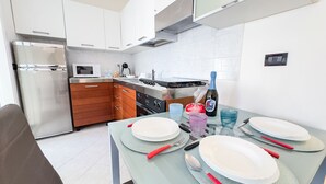 Een moderne keuken met witte kasten, een roestvrijstalen koelkast, een magnetron en een gasfornuis. Er staat een eettafel gedekt met borden, bestek en een fles wijn.