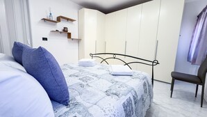 Een slaapkamer met een bed, kussens, een stoel, een kledingkast en planken met spullen.