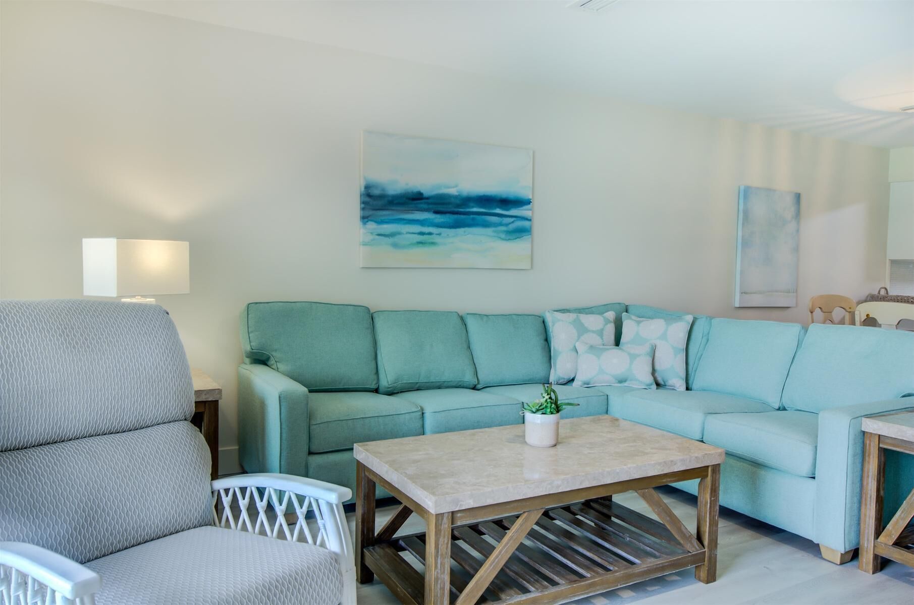 Uma sala de estar com um sofá verde-água, uma poltrona branca, uma mesa de centro de madeira e um quadro com um céu azul.