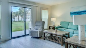 A spacious living room with a sliding glass door leading to a balcony, a grey armchair, a teal sofa, a small table with a lamp, and a painting on the wall. - 2C, Stay Salty (Miramar Beach)