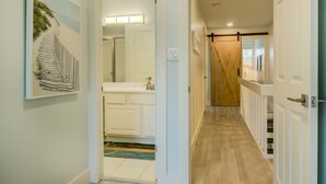 A hallway with a wooden floor, white walls, and a door leading to a bathroom with a mirror and a sink. - 2C, Stay Salty (Miramar Beach)