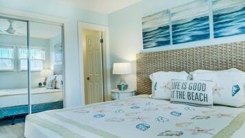 A bedroom with a bed, a nightstand, a lamp, a mirror, and a wall with a beach-themed artwork.