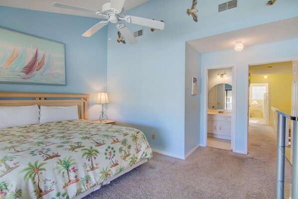 A bedroom with a bed, a ceiling fan, a nightstand, a lamp, a painting, and a hallway leading to a bathroom. - 3 A, Three Bedroom Townhome (Miramar Beach)