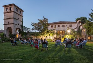 An outdoor gathering with people seated on chairs in a grassy area, a historic building with a tower in the background, and a stage with a performer. - Room-double Room Between Padua and Chiog (Brugine)