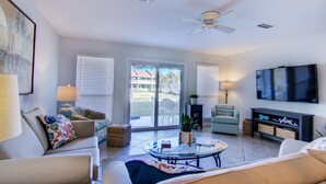 A living room with a TV, a ceiling fan, and a large window. - 2B, Fore Shore! (Miramar Beach)