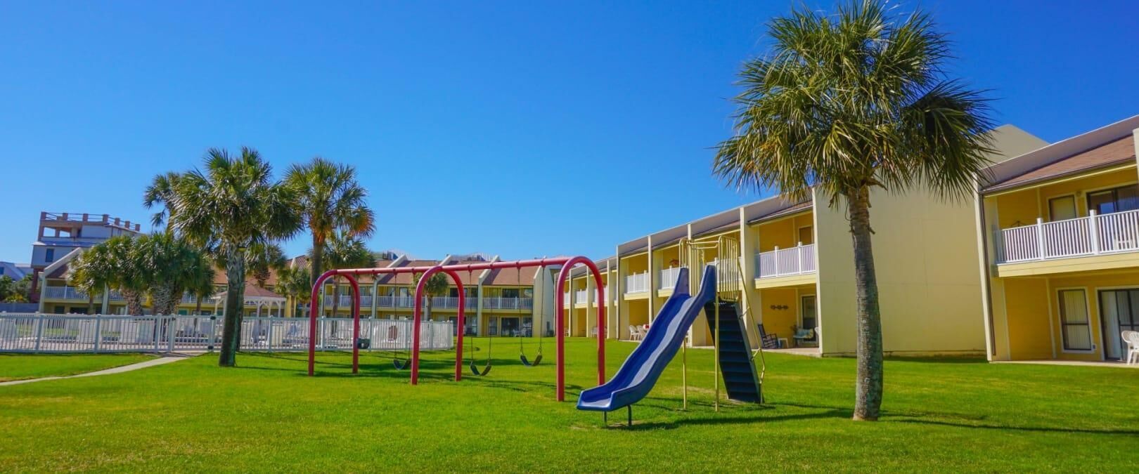 A playground with swings and a slide, surrounded by palm trees and a building with balconies.