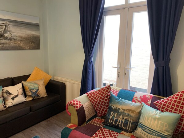 Een woonkamer met een bank en fauteuils, een schilderij aan de muur en uitzicht door een raam met gordijnen.