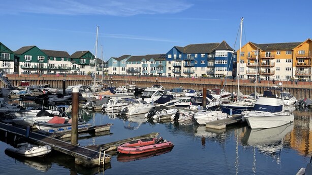 Un port de plaisance où sont amarrés différents types de bateaux, une jetée en bois et, au fond, des bâtiments aux couleurs vives.